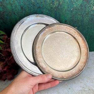 May include: Two silver-colored plates with starburst patterns on the rims. One plate is larger and brighter, while the other shows signs of age with a slightly tarnished appearance. The plates are round and have a classic design.