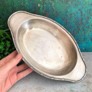 Puede incluir: Una fuente de servir ovalada de tono plateado con un interior liso y pulido y un borde ancho y plano. El borde exterior presenta un patrón decorativo repetitivo de cruces que rodea todo el perímetro del recipiente de metal.