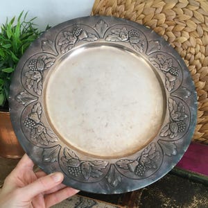 Op de afbeelding: Een ronde zilverkleurige metalen onderbord met een brede, in reliëf gemaakte rand versierd met een herhalend patroon van eikels, eikenbladeren en gestructureerde bessen. Het midden van het bord is glad en rond met een licht aangeslagen afwerking.