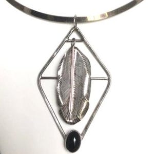 May include: A silver-toned necklace featuring a pendant with a feather design inside a diamond-shaped frame. A small black oval stone is at the bottom of the pendant. The necklace has a simple, thin band.