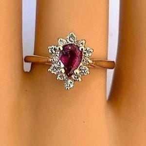 May include: A gold ring featuring a pear-shaped, deep red ruby gemstone surrounded by a halo of small, clear diamonds. The ring's band is a simple, polished gold. The ring is shown on a finger.