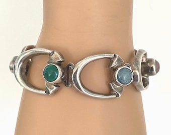 May include: Silver-toned bracelet featuring a series of curved links, each adorned with a central gemstone. The gemstones are a mix of green, blue, and a reddish-brown color. The bracelet is displayed on a wrist.