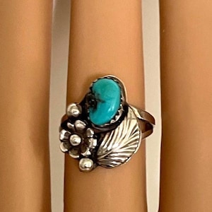 May include: A silver ring featuring a turquoise stone, surrounded by floral and leaf designs. The ring's turquoise stone is oval-shaped and set in a bezel setting. The ring has a vintage or Southwestern style.