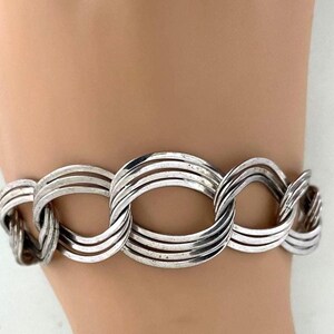 May include: A silver-toned bracelet featuring a series of interconnected oval links. Each link is composed of multiple parallel bands, creating a textured, layered effect. The bracelet is designed to be worn on the wrist.