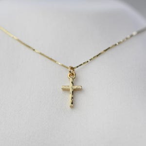 May include: A gold-colored cross pendant necklace. The cross has a textured surface and hangs from a delicate chain. The necklace is displayed against a white background, highlighting the jewelry's details.