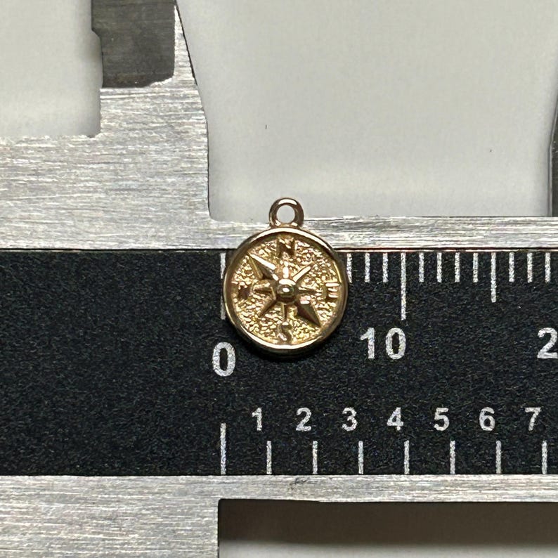 K&ouml;nnte beinhalten: Ein goldfarbener Kompass-Anh&auml;nger mit detailliertem Design. Der Kompass hat einen Durchmesser von etwa 1 cm und eine &Ouml;se oben zum Anbringen an einer Halskette oder einem Armband.