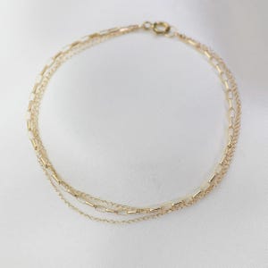 May include: A delicate gold bracelet with three strands of chains. The top strand has rectangular links, the middle has smaller rectangular links, and the bottom is a fine chain. The bracelet is secured with a small clasp. The background is white.