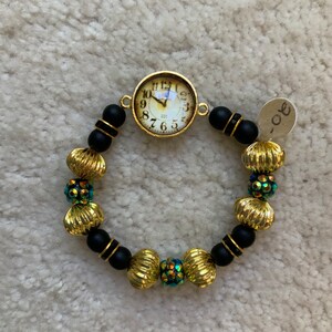 Puede incluir: Un brazalete de cuentas negras y doradas con un dije de esfera de reloj. Las cuentas son redondas y tienen una superficie texturizada. La esfera del reloj es blanca con números y manecillas negros.