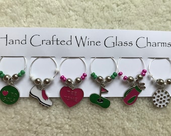 May include: Set of six wine glass charms with golf-themed designs. The charms feature a golf ball, a golf club, a golf bag, a golf shoe, a heart with "Tee Time" written on it, and a heart with "I Love Golf" written on it.