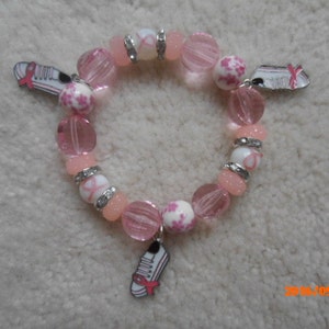 Könnte beinhalten: Ein rosa Perlenarmband mit Charms. Das Armband besteht aus verschiedenen Rosatönen, darunter transparente, opake und gemusterte Perlen. Silberfarbene Abstandshalter und Charms in Form von Laufschuhen mit rosa Bändern sind ebenfalls enthalten.