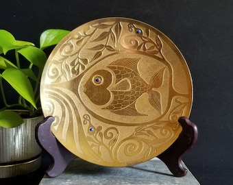 Op de afbeelding: Een gouden metalen bord met een reliëfdesign van een vis en draaiende patronen. De vis heeft blauwe ogen en is omringd door gestileerde planten. Het bord staat op een houten standaard.