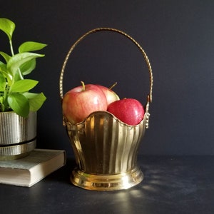 Peut inclure: Un panier en laiton avec une poignée, rempli de trois pommes rouges. Le panier a un bord festonné et une base ronde. Une plante verte dans un pot doré est à gauche du panier.