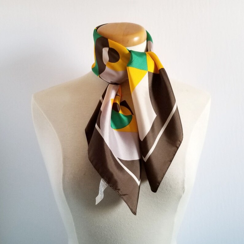 May include: A colorful silk scarf with a geometric pattern. The scarf features a combination of brown, white, yellow, and green shapes. The scarf is draped around a mannequin's neck, showcasing its design and style.