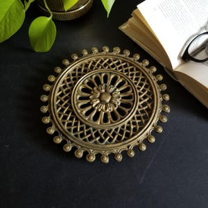 May include: A round, brass trivet with a decorative, intricate design. The trivet has a circular, openwork centre with a floral pattern and a beaded outer edge.