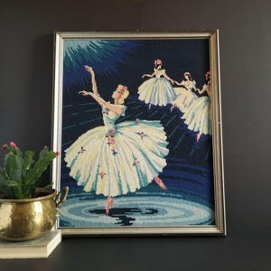 May include: Framed needlepoint artwork depicting three ballerinas in white tutus dancing on a blue background. The ballerinas are in various poses, with one in the foreground and two in the background. The artwork is framed in a silver frame.