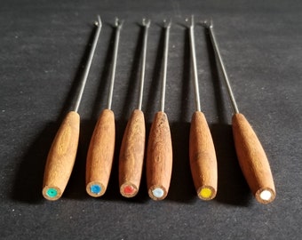 Op de afbeelding: Set van zes fondue vorken met houten handvatten. De vorken hebben zilveren metalen tanden en gekleurde stippen aan de uiteinden van de handvatten. De kleuren zijn groen, blauw, rood, wit, geel en wit.