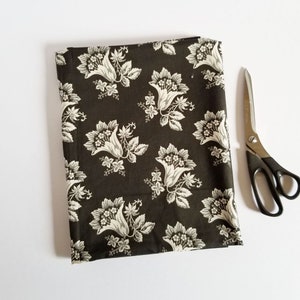Op de afbeelding: Een zwarte stof met een wit bloemenpatroon. De stof heeft een herhalend patroon van gestileerde bloemen met een klokvorm.