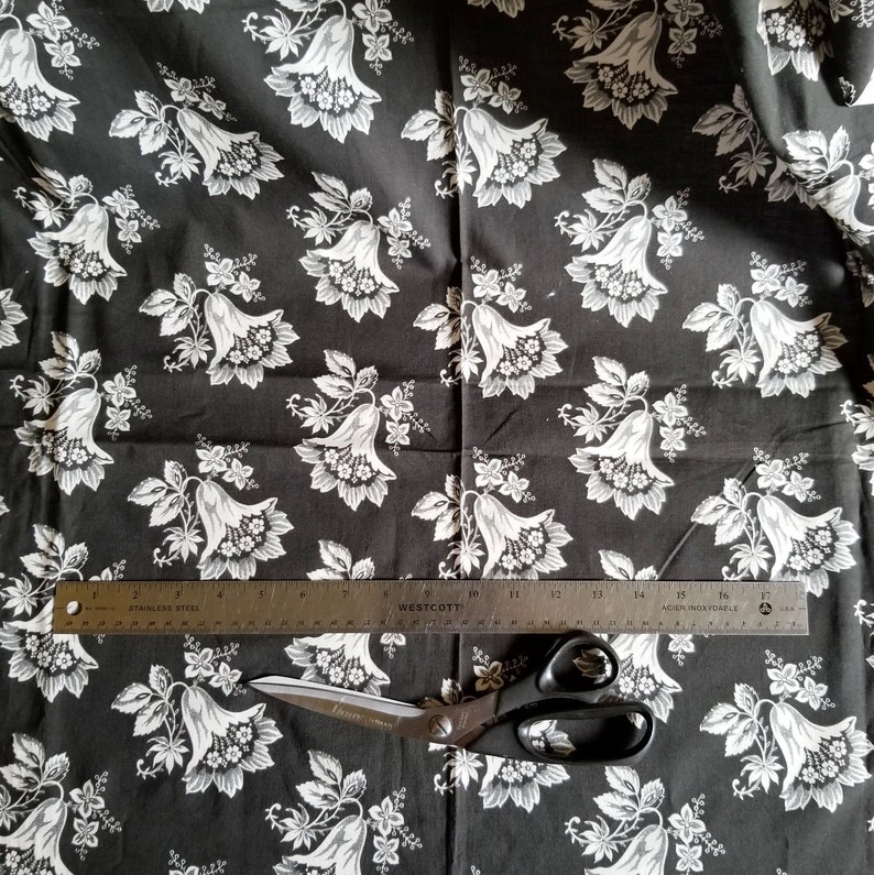 Op de afbeelding: Zwarte stof met een wit bloemenpatroon van klokvormige bloemen. Een liniaal en een schaar liggen op de stof.