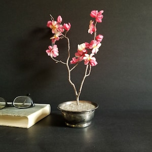 May include: A small, silver-coloured planter with a delicate, twig-like branch with pink and white blossoms. The planter is filled with small, white beads.