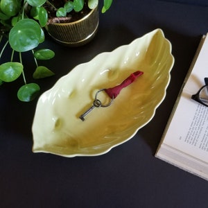 Peut inclure: Un plat en céramique jaune en forme de feuille avec une clé et un ruban rouge à l'intérieur. Le plat est sur une surface noire.