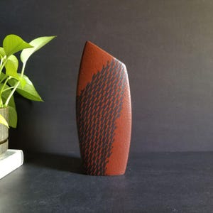 May include: A red ceramic vase with a black abstract pattern. The vase is tall and narrow, with a rounded base and a pointed top.
