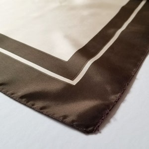 May include: A silk scarf with a cream-colored center and a dark brown border. The border is accented with a thin white stripe. The scarf's corner is visible, showcasing the fabric's texture and the stitched edge.