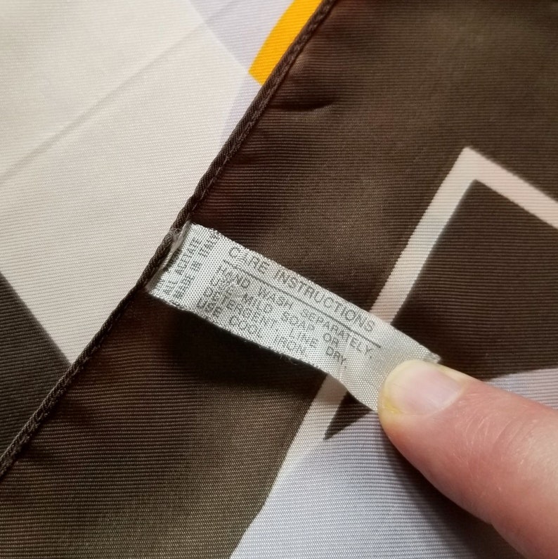 May include: A silk scarf with a geometric pattern in brown, white, and yellow. The scarf has a tag that reads "ALL ACETATE MADE IN ITALY" and care instructions: "HAND WASH SEPARATELY, USE MILD SOAP OR DETERGENT. LINE DRY. USE COOL IRON."