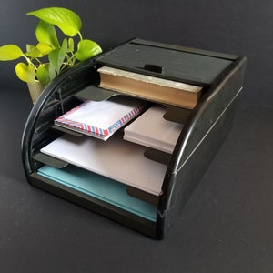 Peut inclure: Organiseur de bureau en plastique noir avec quatre compartiments. Le compartiment supérieur a un couvercle. L'organisateur est rempli d'enveloppes, de papier et d'un livre.