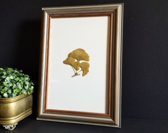 Op de afbeelding: Een ingelijst gedrukt ginkgo-blad kunstwerk met een witte passe-partout en een zilveren lijst. Het blad is lichtgeel van kleur en is plat gedrukt tegen de witte passe-partout.