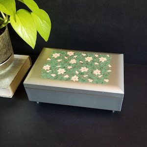 May include: A rectangular gray jewelry box with a silver lid. The lid is decorated with a floral pattern of white flowers and green leaves. The box is on a black surface.