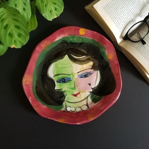 May include: A ceramic plate featuring a hand-painted design of a woman's face. The woman has black hair, green and pink skin, and blue eyes. The plate is decorated with a red, green, and yellow border.