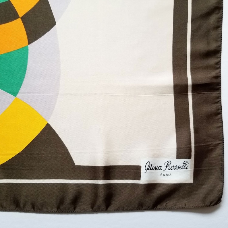 May include: A square silk scarf with a geometric design. The scarf features a cream-colored background with a brown border and accents. The design includes yellow, green, and black shapes. The label reads "Olivia Rosselli ROMA."