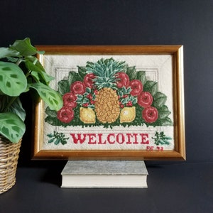 May include: Cross-stitch picture in a gold frame, with a pineapple, red roses, and the word "WELCOME". The artwork has a neutral background. The frame is made of wood.