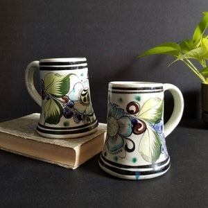 May include: Two white ceramic mugs with black trim and colourful floral designs. The mugs have a flared base and a handle.