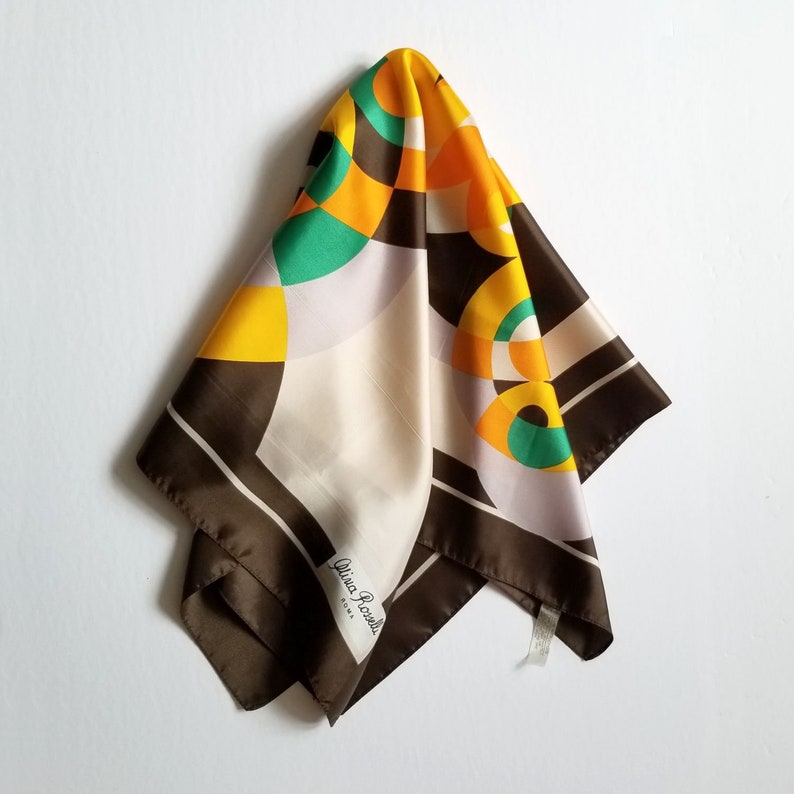 May include: A silk scarf with a geometric pattern in shades of brown, yellow, green, and cream. The scarf has a dark brown border and the text "Olivia Rossi Roma" is visible. The scarf is hanging against a white background.