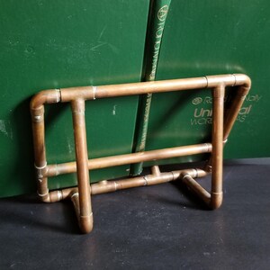May include: A copper pipe bookend with two books resting on it. The books are green and have gold lettering on the spine. The bookend is made of copper pipe and has a simple design.