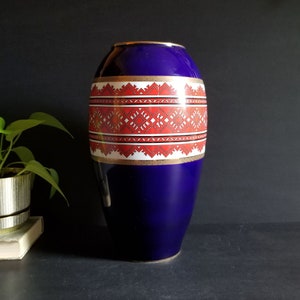 May include: A tall, cobalt blue ceramic vase with a geometric pattern in red, white, and gold. The vase has a rounded body and a narrow opening.