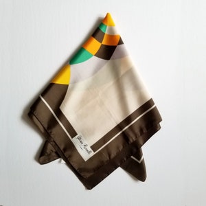 May include: A folded silk scarf with a geometric design. The scarf features a brown border with white stripes, a cream-colored center, and colorful geometric shapes in yellow, green, and orange. The label reads "Gina Roselli ROMA".
