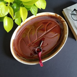 May include: A brown and white oval ceramic dish with a raised leaf design. A silver key and a red ribbon are on the dish.