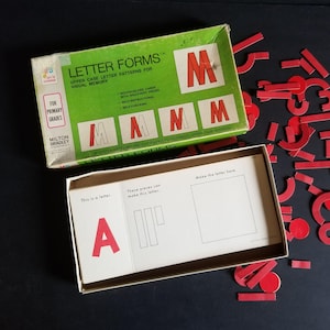 May include: A vintage educational toy for primary grades called "Letter Forms" by Milton Bradley. The box features a green background with red uppercase letters "M" and the text "Letter Forms" in white. The box is open, revealing a white tray with red uppercase letters "A" and "M" and instructions for assembling the letters.