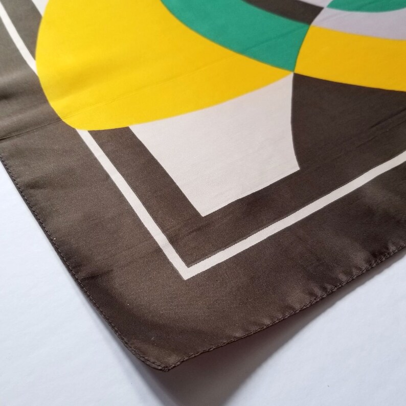 May include: A square silk scarf with a geometric design. The scarf features a brown border with a white inner border. The interior design includes yellow, green, white, and gray shapes. The scarf is folded, showing a corner.