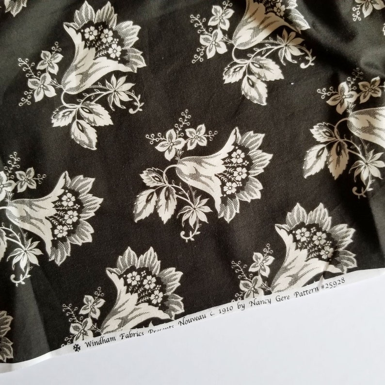 Op de afbeelding: Zwart-witte stof met een herhalend patroon van gestileerde bloemen. De stof is gelabeld "Windham Fabrics  *Nouveau c. 1910 by Nancy Gere Pattern #25928"