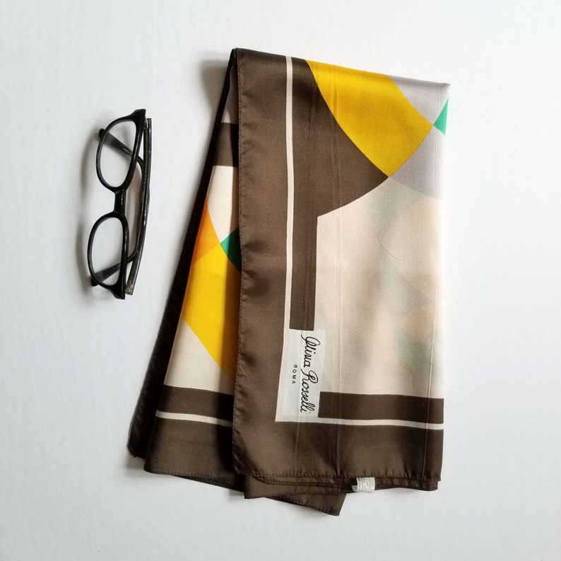 May include: A folded silk scarf with a geometric design in yellow, green, and cream, framed by a brown border. Black eyeglasses are positioned to the left of the scarf. The scarf has a tag that reads "Alisa Rossi ROMA."