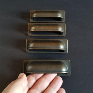 May include: Four antique bronze cabinet pulls with a rounded rectangular shape and a slightly curved top. The pulls are arranged vertically in a row.