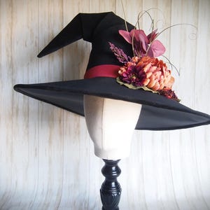 Pode incluir: Um chapéu de bruxa preto com aba larga e coroa pontuda, adornado com uma faixa vermelha e um aglomerado de flores artificiais em tons de laranja, bordô e rosa. O chapéu é exibido em uma cabeça de manequim, sugerindo uma fantasia ou acessório.