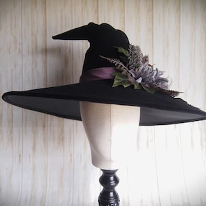 Pode incluir: Um chapéu de bruxa preto com aba larga e copa pontiaguda, decorado com uma fita roxa e detalhes florais. O chapéu é exibido em uma cabeça de manequim contra um fundo de madeira clara.