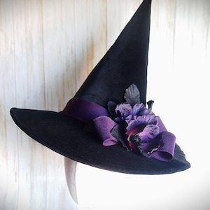 Pode incluir: Um chapéu de bruxa preto com aba larga e coroa alta e pontiaguda. O chapéu é adornado com uma fita roxa e um aglomerado de flores de tecido roxas e pretas. Acessório de Halloween ou fantasia.