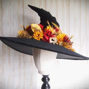 Pode incluir: Um chapéu de bruxa preto com aba larga, adornado com um arranjo vibrante de flores coloridas de outono. O chapéu é exibido em uma cabeça de manequim, mostrando sua forma e design. As flores incluem flores vermelhas, amarelas e brancas.