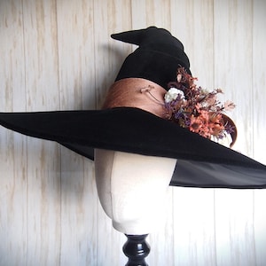 Pode incluir: Um chapéu de bruxa preto com aba larga e copa pontuda. O chapéu apresenta uma faixa marrom e é adornado com flores secas em tons de laranja, roxo e branco. O chapéu é exibido em uma cabeça de manequim.