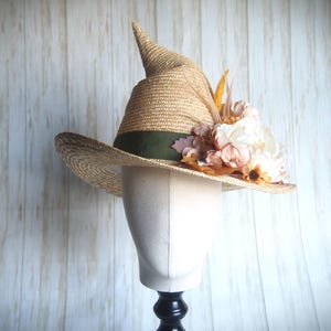 May include: A tan straw witch hat featuring a pointed crown and a wide brim. The hat is accented with a dark olive green ribbon band and a cluster of artificial flowers in shades of cream, peach, and brown, along with dried foliage elements.